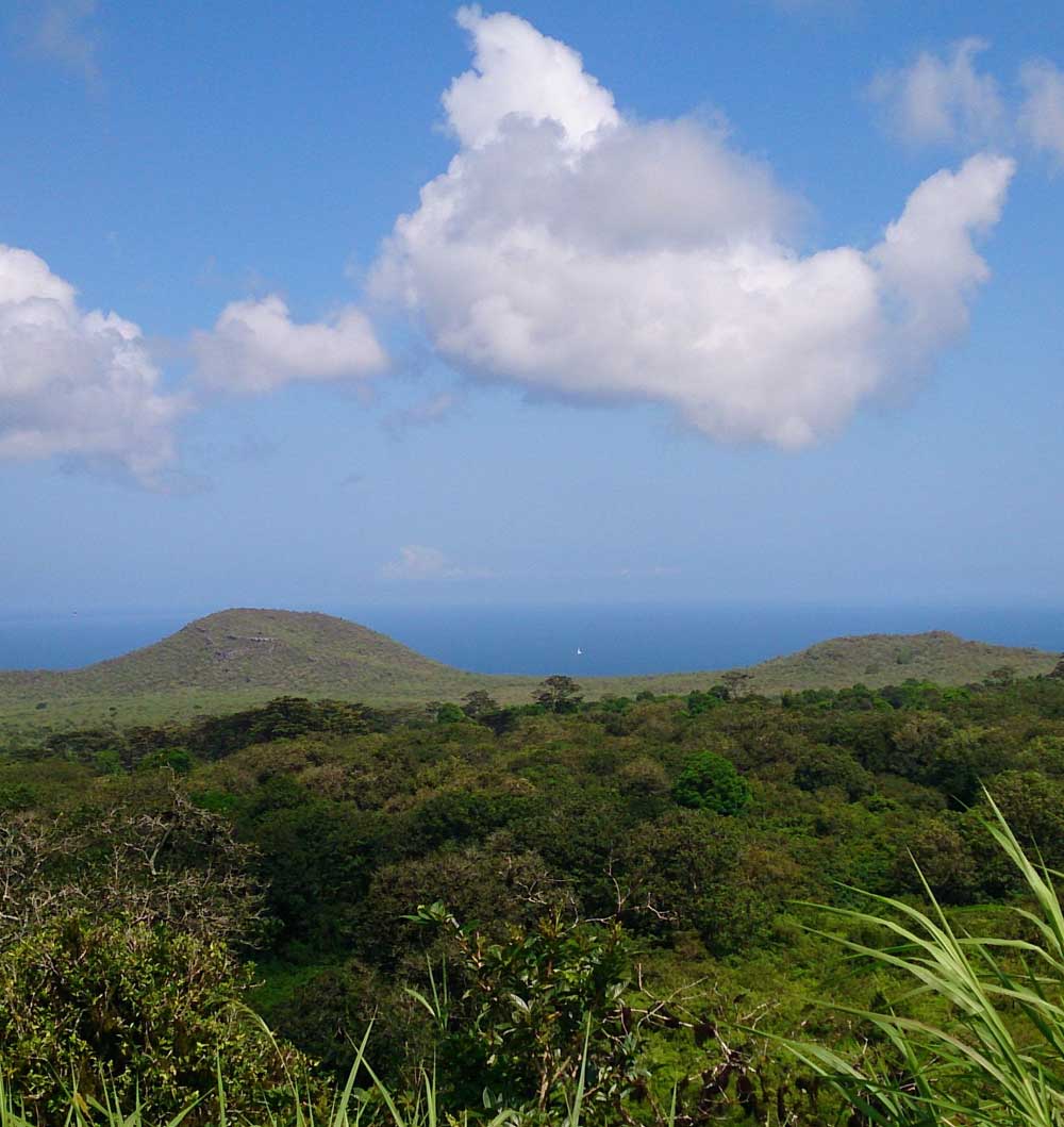 Grüne Hügel auf der Insel San Cristobal mit weissen Wolken am Himmel und dem Meer im Hintergrund.