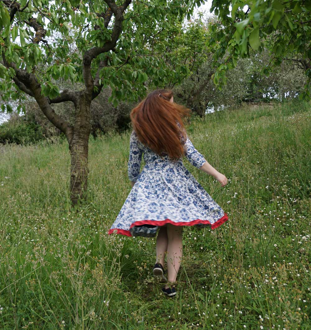 Energietherapeutin Manuela Krah tanzt in einem blau-weissen Sommerkleid mit markantem rotem Saum auf einer Wiese zwischen Bäumen hindurch,