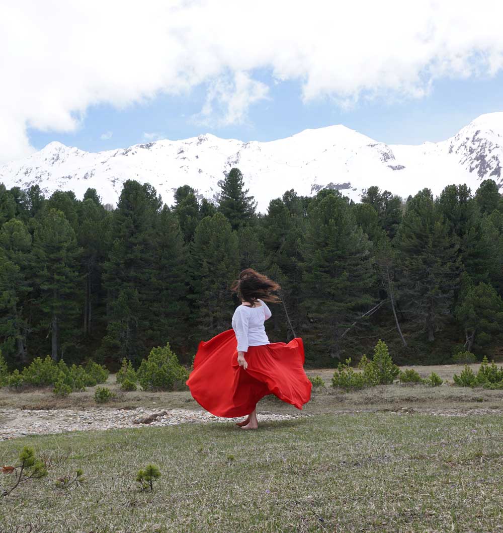 Energietherapeutin Manuela Krah tanzt in einem roten Rock und weisser Bluse neben einem Bach. Im Hintergrund sind dunkelgrüne Tannen und dahinter weisse Schneeberge zu sehen.