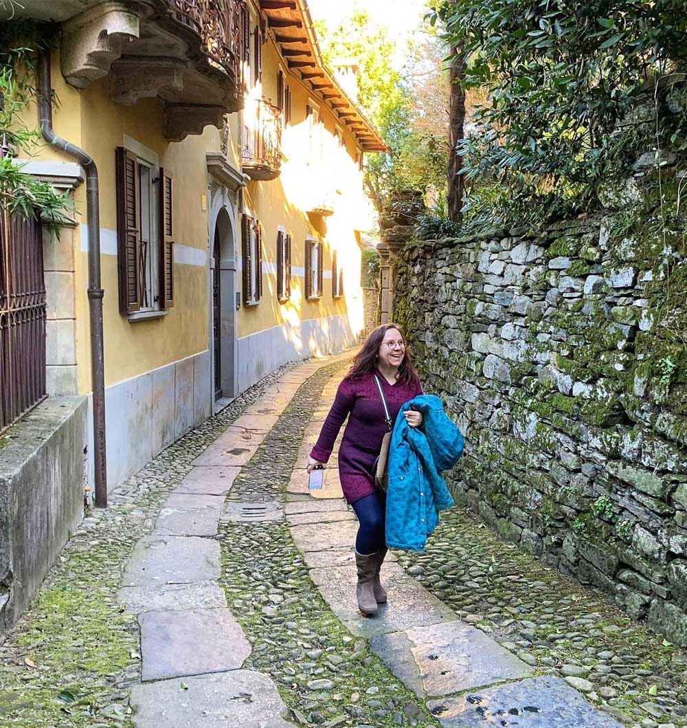 Die Energietherapeutin Manuela Krah schreitet über einen Weg mit Kopfsteinpflaster in Isola San Giulio Lago d’arta, Italien. Sie trägt eine hellblaue Jacke über dem Ärmel.