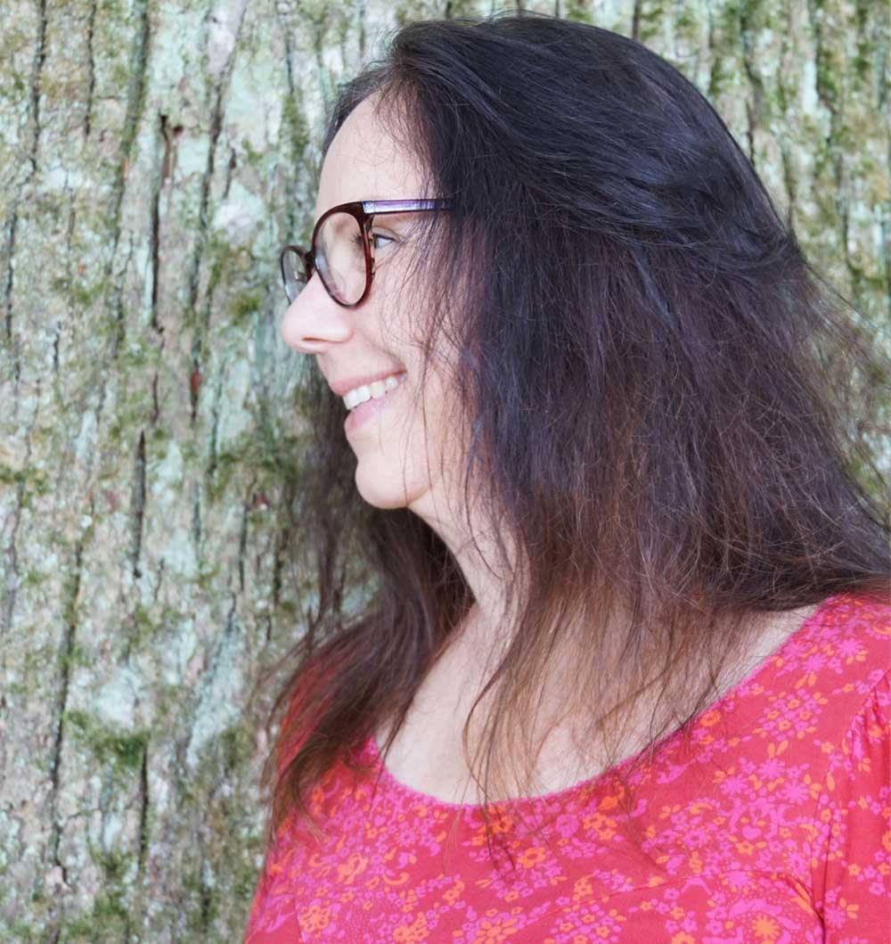 Seitenaufnahme der Energietherapeutin Manuela Krah. Sie steht vor einem Baum, hat schulterlange dunkelbraune Haare und trägt ein rotes Shirt mit Ornament-Muster.