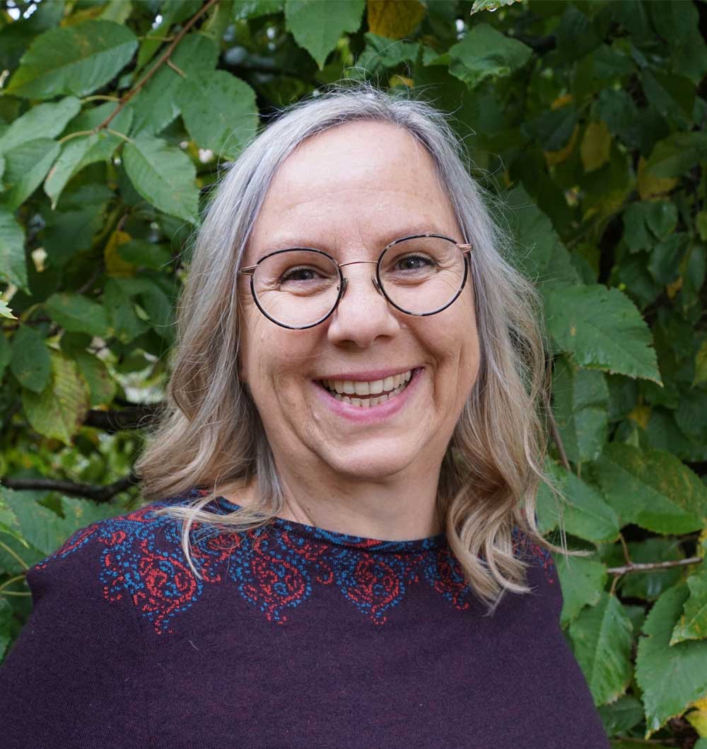 Energietherapeutin Manuela Krah. Sie steht vor einem grünen Busch. Sie hat schulterlanges, graues Haar und trägt eine runde Brille und einen violetten Pullover.