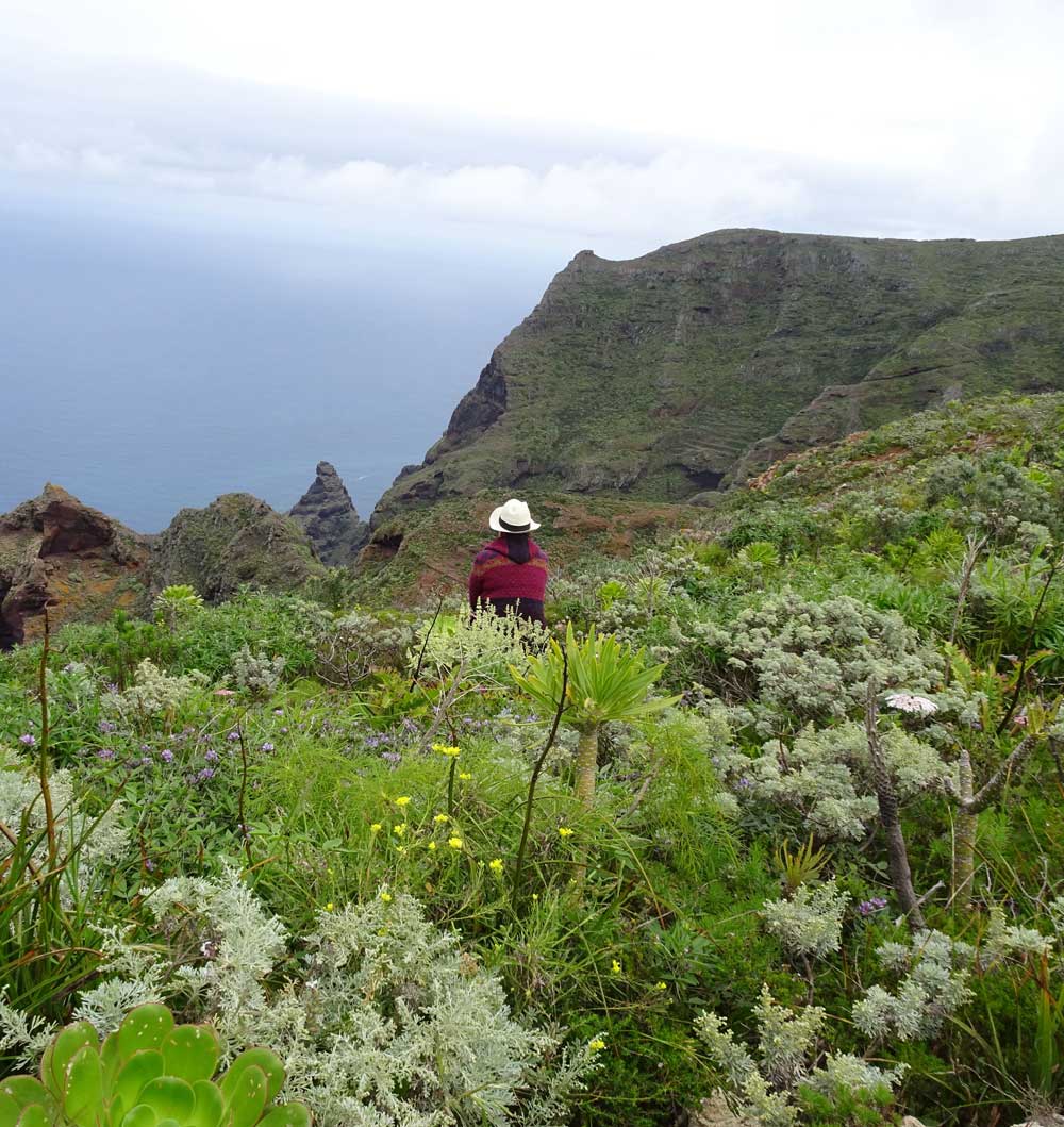 Die Energietherapeutin Manuela Krah sitzt auf einer grünen, dichtbewachsenen Klippe und schaut aufs Meer hinaus. Sie trägt einen weissen Panama Hut und einen roten Umhang, wie er typisch für die indigenen Völker Südamerikas ist.