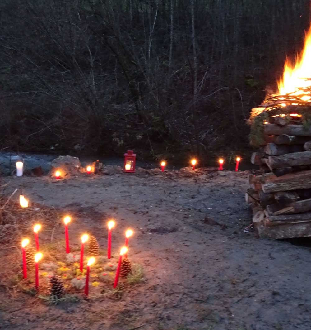Ein sorgfältig aufgebauter Holzhaufen brennt. Zwischen dem Holz befinden sich die Steine für die Schwitzhüttenzeremonie. Links im Vordergrund ist ein Kreis mit brennenden, roten Kerzen zu sehen. Im Hintergrund ist der Wegrand mit brennenden Kerzen und Laternen markiert.