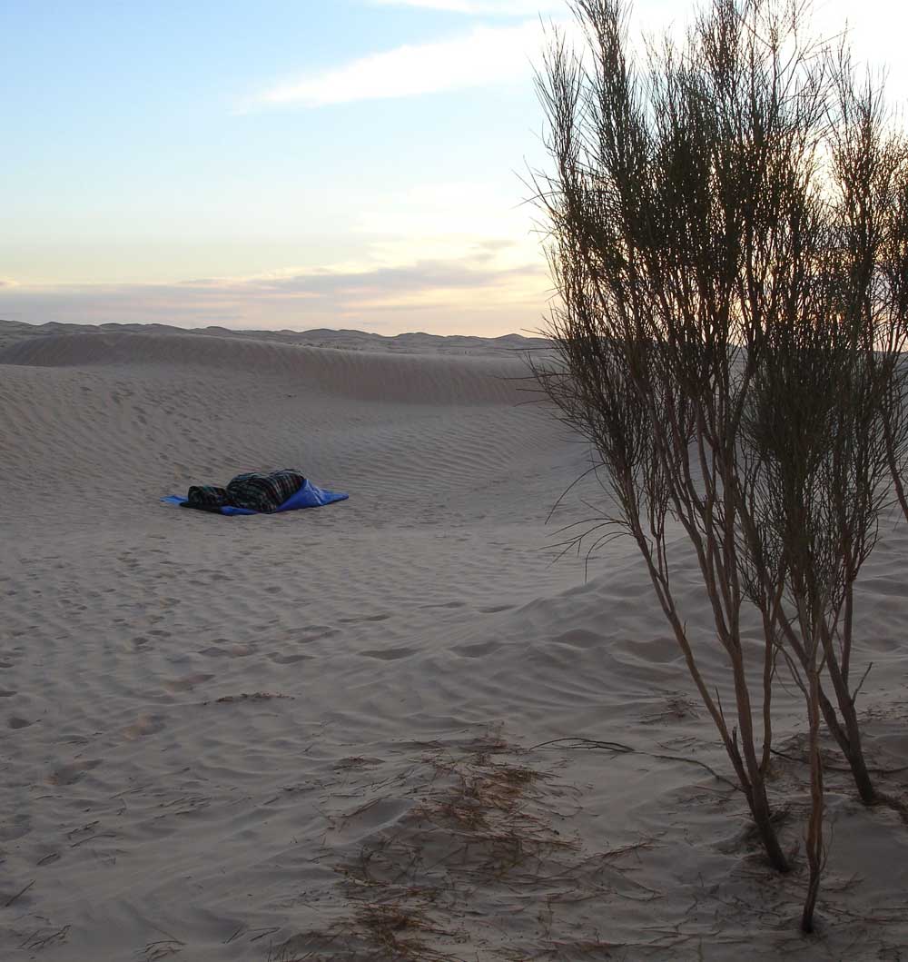 Die Energietherapeutin Manuela Krah liegt in einem blauen Schlafsack in der Sahara. Im Hintergrund links sind Hügelzüge; im Vordergrund rechts Sträucher zu sehen.