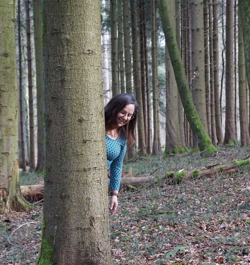 Energietherapeutin Manuela Krah steht im Wald und blickt hinter einem Baum hervor.