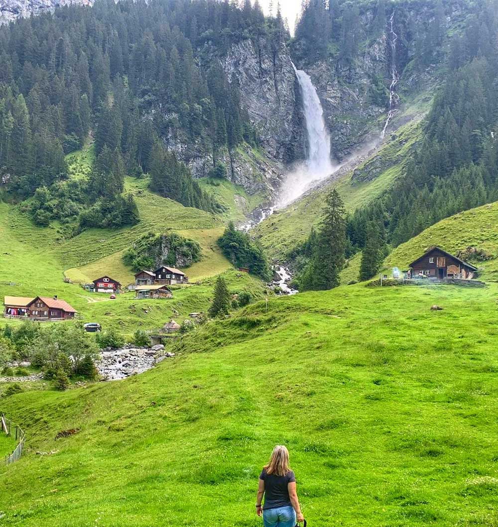 Manuela steht am Fusse eines kleinen Bergdorfes und schaut auf einen Wasserfall.