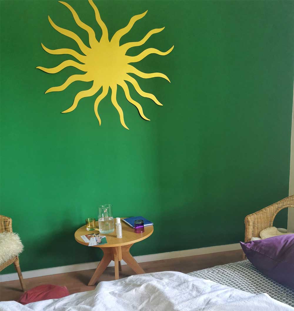 Neben einer Liege mit 2 dünnen Decken darauf steht ein Holztischchen mit 2 Stühlen. Dahinter an der grünen Wand hängt eine gelbe Sonne.
