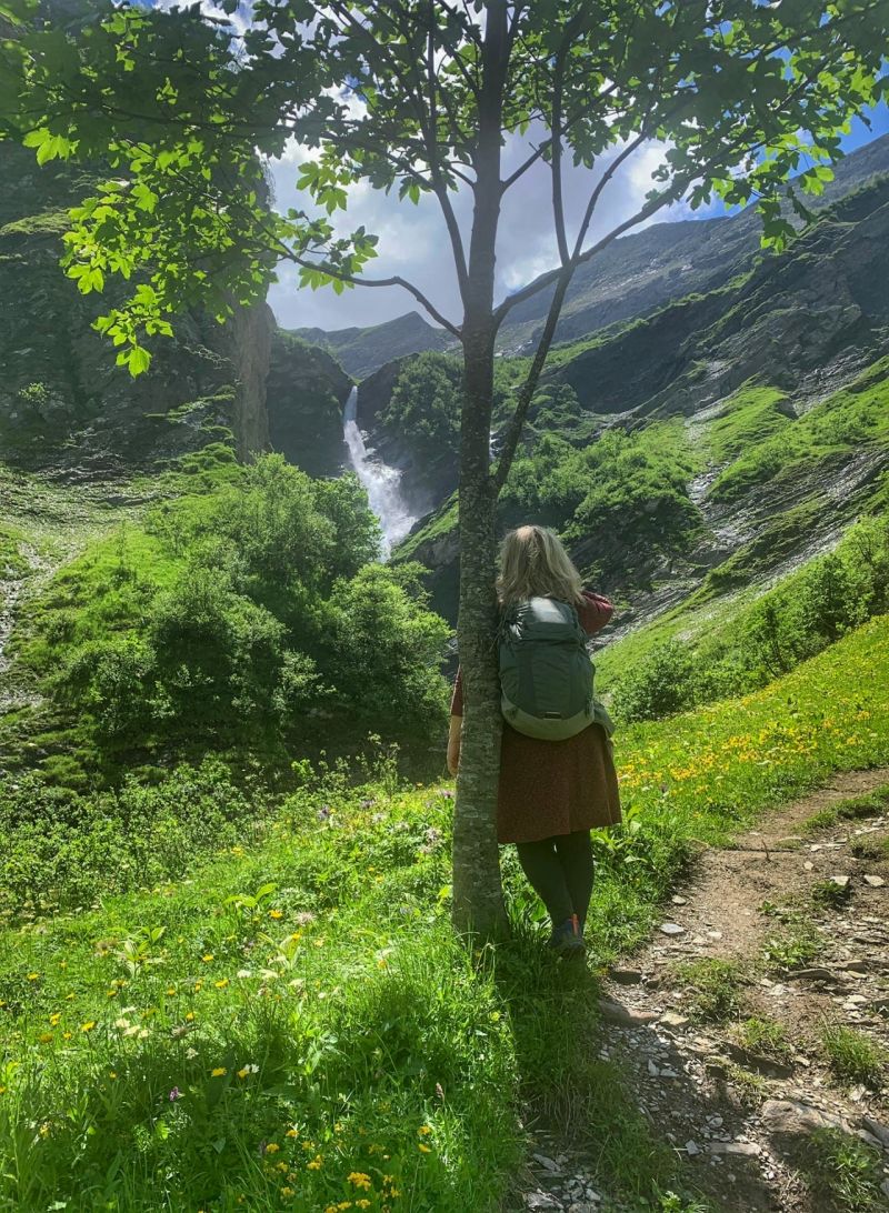Manuela Krah steht mit einer Sonnenbrille vor Wald und Hügel. Der Wald geht um den Caumasee herum. 