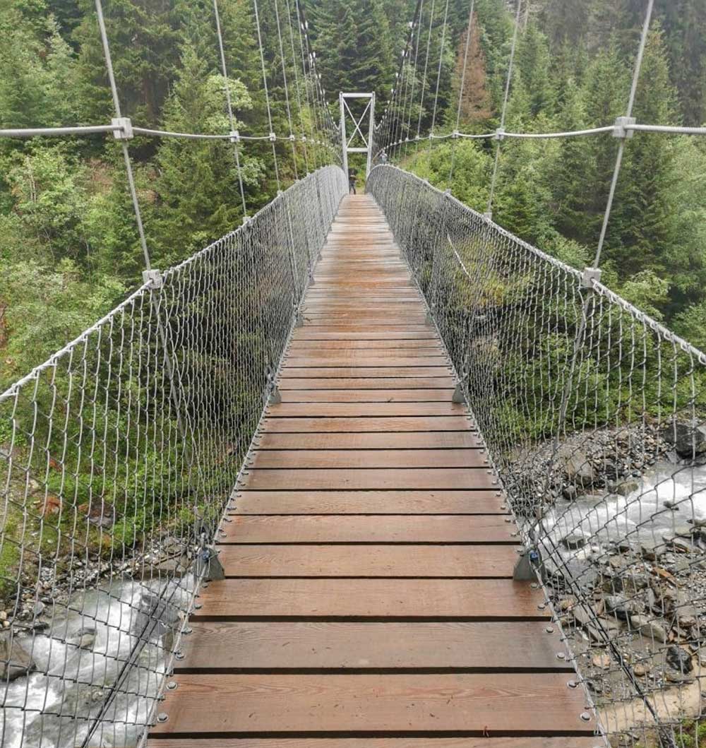 Eine Holzbrücke über einem steinigen Bach führt auf der anderen Seite in einen grünen Wald.