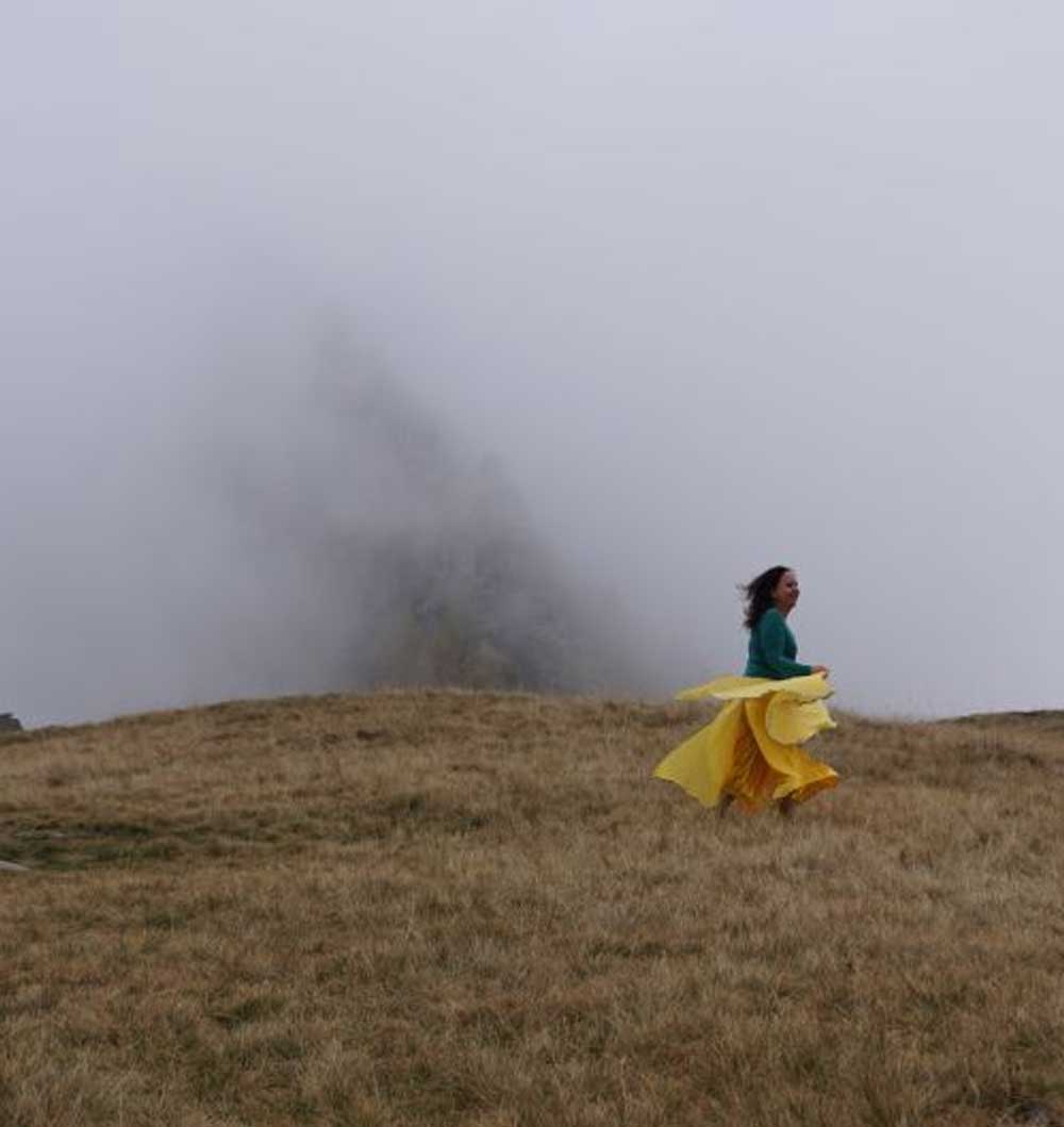 Manuela Krah läuft in einem wallenden gelben Kleid über eine trockene Wiese. Im Hintergrund ist Nebel zu sehen.